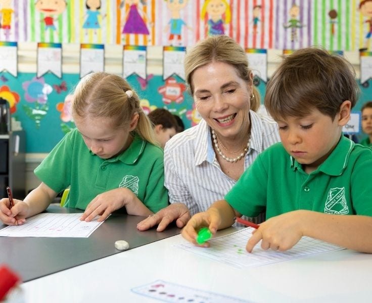 girl and boy student working on their writing whilst the Teacher helps them in brightly coloured classroom